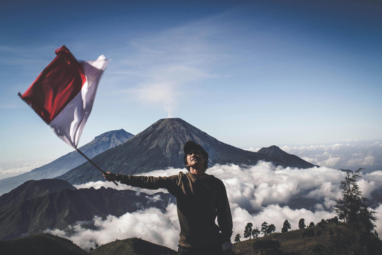 A hiker waves the Indonesian flag atop a scenic mountain, surrounded by clouds.