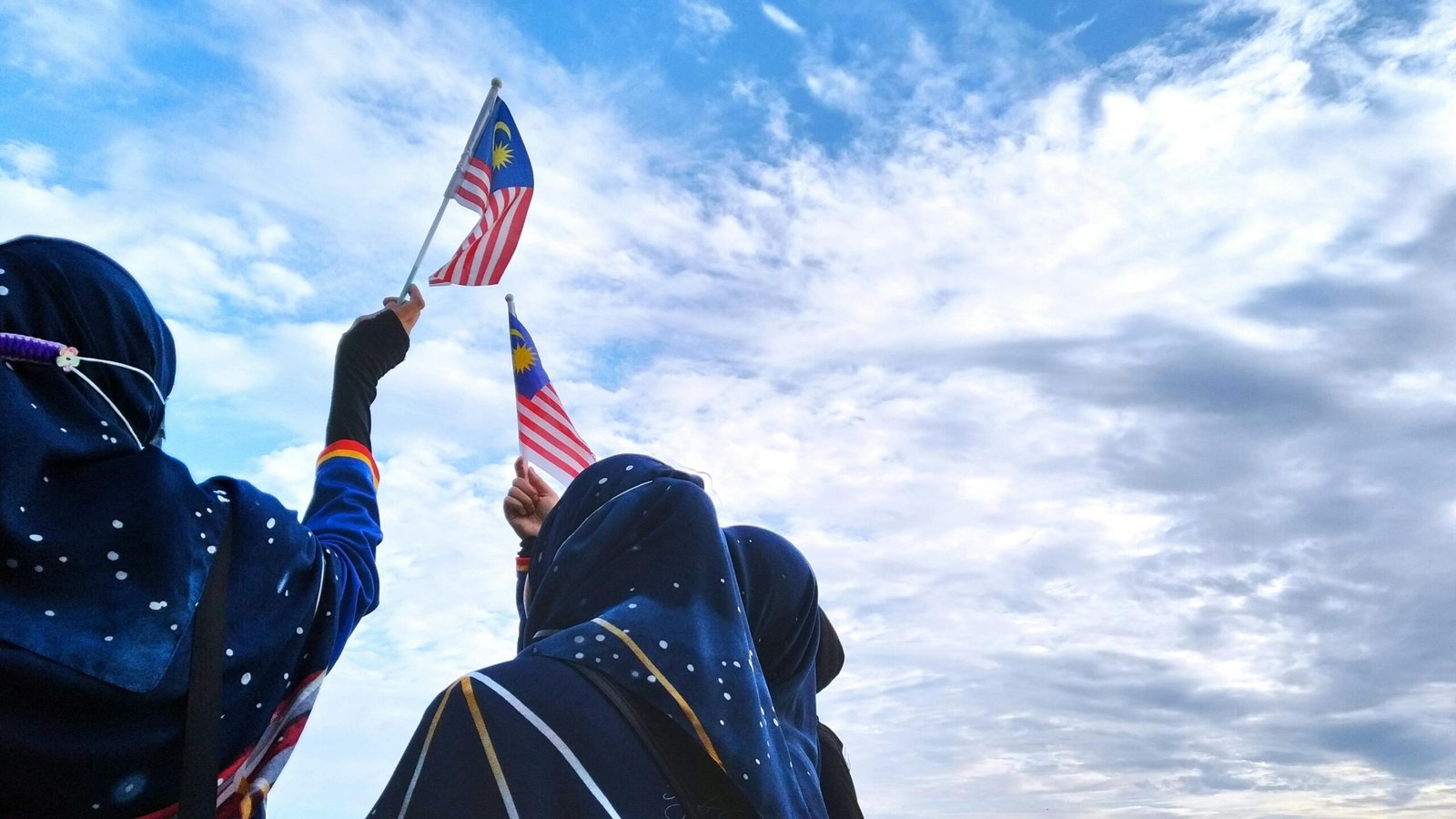 Malaysian women celebrating patriotism, waving flags against a clear sky.