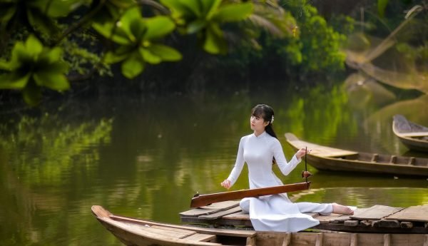 Woman in white dress playing Dan Bau on a boat amid lush greenery.